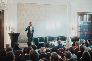 Elegant gekleidetes Publikum lauscht einem Vortragenden auf einer Bühne in einem historischen Saal.