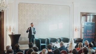 Elegant gekleidetes Publikum lauscht einem Vortragenden auf einer Bühne in einem historischen Saal.