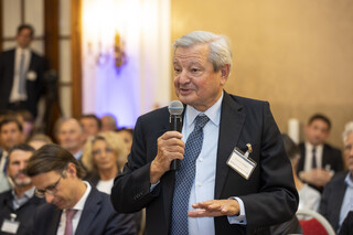 Mann mit grauem Haar spricht in ein Mikrofon, das er in der Hand hält und bewegt die andere Hand. Er trägt ein schwarzes Sakko, ein hellblaues Hemd und eine blau-karierte Krawatte.