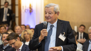 Mann mit grauem Haar spricht in ein Mikrofon, das er in der Hand hält und bewegt die andere Hand. Er trägt ein schwarzes Sakko, ein hellblaues Hemd und eine blau-karierte Krawatte.