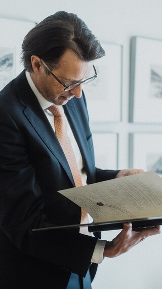 Ein Herr in Geschäftskleidung blättert vor einer mit Gemälden bestückten Wand in einem historischen Buch.