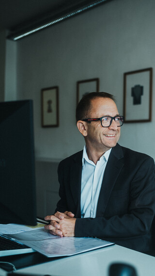 Ein lächelnder Herr in Geschäftskleidung sitzt an einem Schreibtisch und blickt zur Seite.