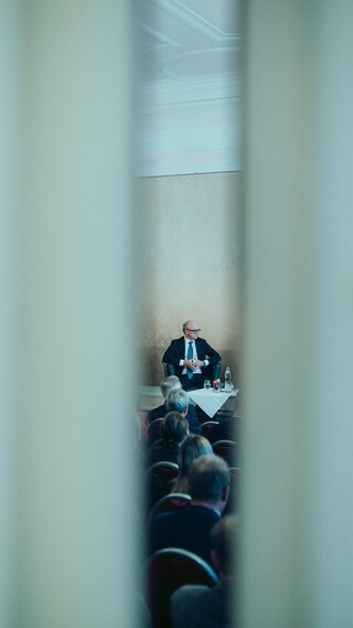 Blick durch einen Schlitz auf eine Bühne mit einer Podiumsdiskussion und deren Publikum.