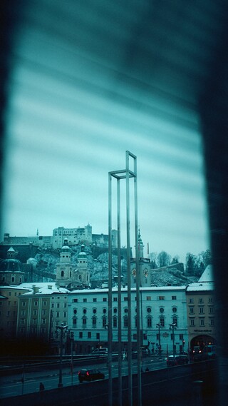 Blick durch ein Fenster auf einen Sessel mit überdimensional langen Füßen vor dem Hintergrund einer verschneiten Altstadt.
