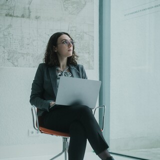 Eine Frau mit Brille sitzt auf einem Stuhl, auf ihrem Schoß liegt ein Laptop. 