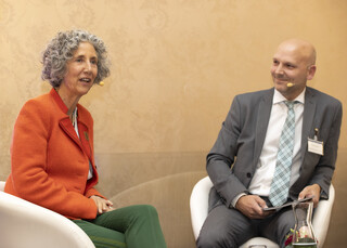 Eine Frau und ein Mann diskutieren am Podium, den Hintergrund ziert eine beige-gemusterte Wand. Die Frau hat gräuliche Locken und trägt einen orange-farbigen Blazer und eine grüne Hose. Der Mann trägt einen grauen Anzug, ein weißes Hemd und eine blau-grau karierte Krawatte.