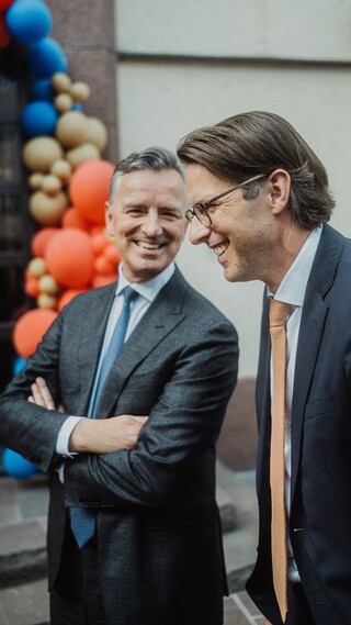 Zwei lachende Herren in Geschäftskleidung stehen vor einem mit Luftballons geschmückten Portal.