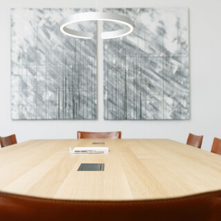 Besprechungsraum mit einem hellen, eckigen Holztisch. Mehrere dunkelbraune Stühle sind darum platziert. Eine graue, rundliche Leuchte hängt von der Dicke über die Mitte des Tisches. Zwei grau-weiße Bilder mit Strichen hängen an der Wand.