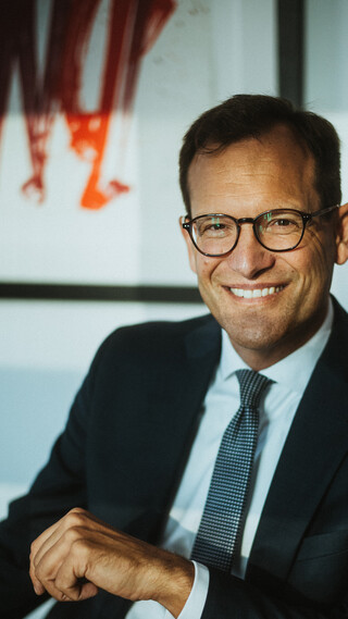 Ein lächelnder Herr in Geschäftskleidung sitzt vor einem Gemälde.