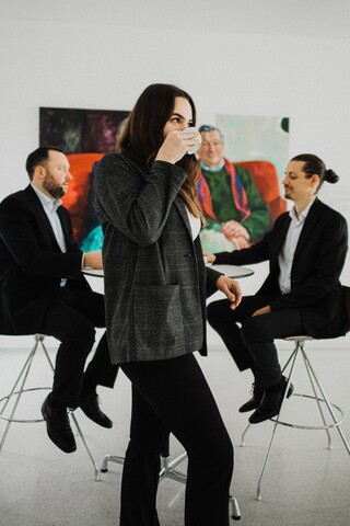 Eine Frau in schwarzem Kostüm steht in einem Raum und trinkt Kaffee.