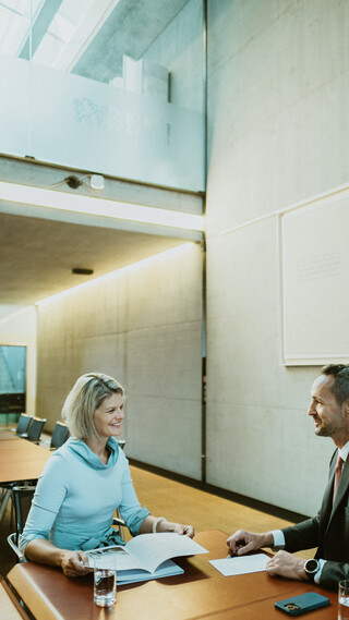 Eine Dame und ein Herr in Geschäftskleidung führen ein Gespräch an einem Tisch in einem modernen Raum.