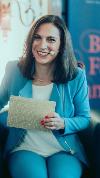 Eine sitzende lächelnde Dame in Geschäftskleidung ist mit Headset und Moderationskarten ausgerüstet.