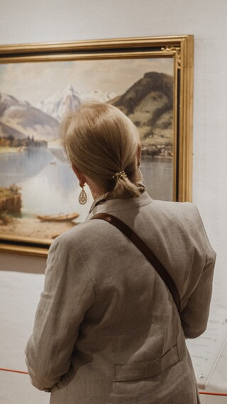 Eine elegant gekleidete Dame blickt auf eine Wand mit Landschaftsgemälden.