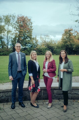 Vier Personen lächeln draußen in die Kamera, mit einer Wiese und Bäumen im Hintergrund.