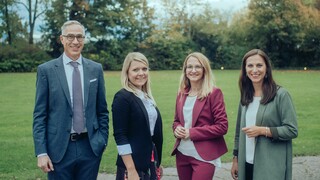 Vier Personen lächeln draußen in die Kamera, mit einer Wiese und Bäumen im Hintergrund.