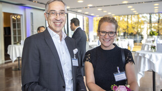 Ein Mann und eine Frau stehen an einem Stehtisch und lächeln in die Kamera. Beide tragen eine Brille.