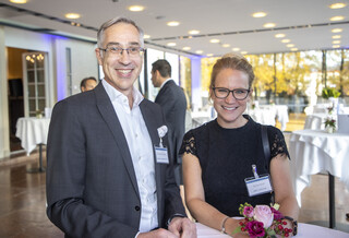 Ein Mann und eine Frau stehen an einem Stehtisch und lächeln in die Kamera. Beide tragen eine Brille.