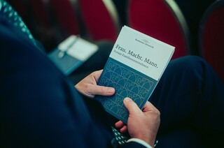 Die Hände eines Mannes sind sichtbar, während er eine Broschüre mit dem Text "Frau. Macht. Mann." beim Forum Familienunternehmen 2024 hält.