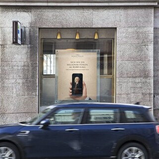 Dunkelblaues Auto (Marke: Mini) fährt an einem Gebäude des Bankhaus Spängler vorbei. Auslagefenster und ein Bankomat-Hinweisschild sind ersichtlich.