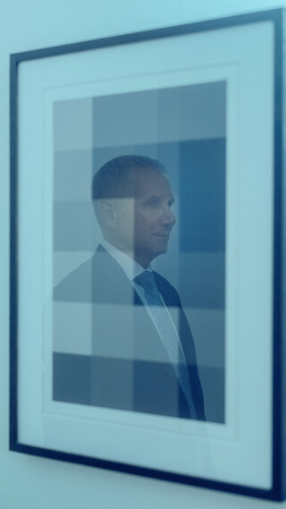 An einer Wand hängt ein Bild mit vielen Vierecken in verschiedenen Grautönen. In der Glasreflexion des Bildes ist ein Mann im Anzug mit weißem Hemd und blauer Krawatte zu sehen.