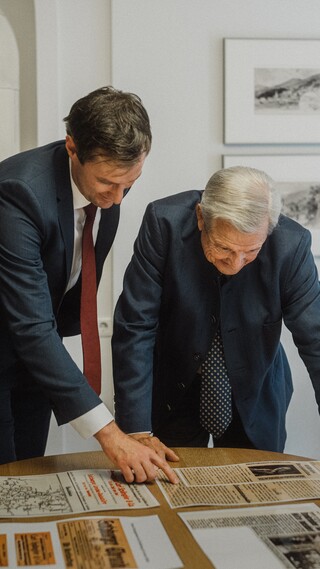 Zwei Herren in Geschäftskleidung lesen historische Zeitungsberichte, die auf einem Tisch liegen.