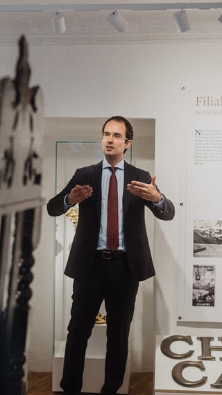 Blick über die Schultern zweier Herren auf einen Vortragenden in Geschäftskleidung.