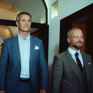 Zwei Herren in Anzug gehen lächelnd einen Gang entlang.