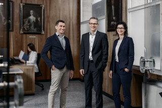 Eine Dame und zwei Herren in Geschäftskleidung stehen lächelnd in einem Büro.