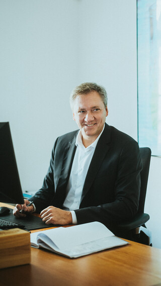 Ein lächelnder Herr in Geschäftskleidung sitzt an einem modernen Schreibtisch.
