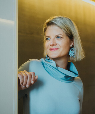 Eine Dame in Kleid steht vor einer Betonwand und blickt lächelnd in die Ferne.