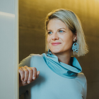 Eine Dame in Kleid steht vor einer Betonwand und blickt lächelnd in die Ferne.