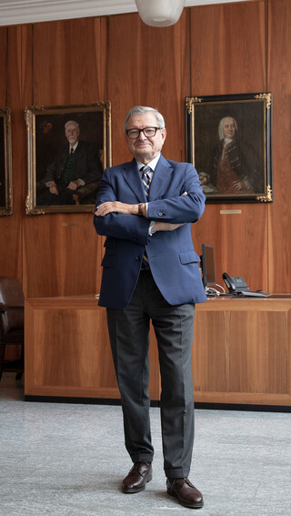 Mann mit Brille, Anzug und verschränkten Händen steht in einem Büro mit dunkler Holzverkleidung. Im Hintergrund sind 4 Gemälde/ Portraits erkennbar.