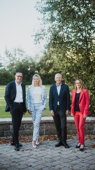Zwei Damen und zwei Herren in Anzug posieren lächelnd vor einem Garten.