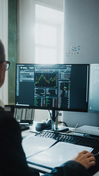 Ein Herr in Geschäftskleidung sitzt vor einem Computerbildschirm mit finanzmathematischen Darstellungen.