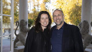 Eine schwarz-gekleidete Frau mit schwarzen, langen Locken und ein dunkel-gekleideter Mann mit Glatze, Bart und Brille am Kopf lächeln. Sie stehen vor einer ins Freie geöffneten Tür.