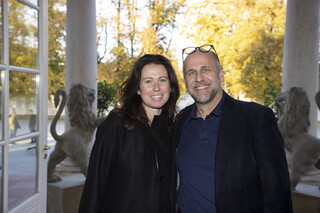 Eine schwarz-gekleidete Frau mit schwarzen, langen Locken und ein dunkel-gekleideter Mann mit Glatze, Bart und Brille am Kopf lächeln. Sie stehen vor einer ins Freie geöffneten Tür.