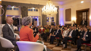 Ein Mann und eine Frau sitzen auf einem Podium in beige-farbigen Stühlen, sie sind von hinten zu sehen. Im Bild zu sehen sind mehrere Teilnehmer, die in Kinobestuhlung dem Vortrag lauschen.