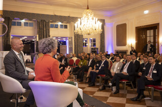 Ein Mann und eine Frau sitzen auf einem Podium in beige-farbigen Stühlen, sie sind von hinten zu sehen. Im Bild zu sehen sind mehrere Teilnehmer, die in Kinobestuhlung dem Vortrag lauschen.
